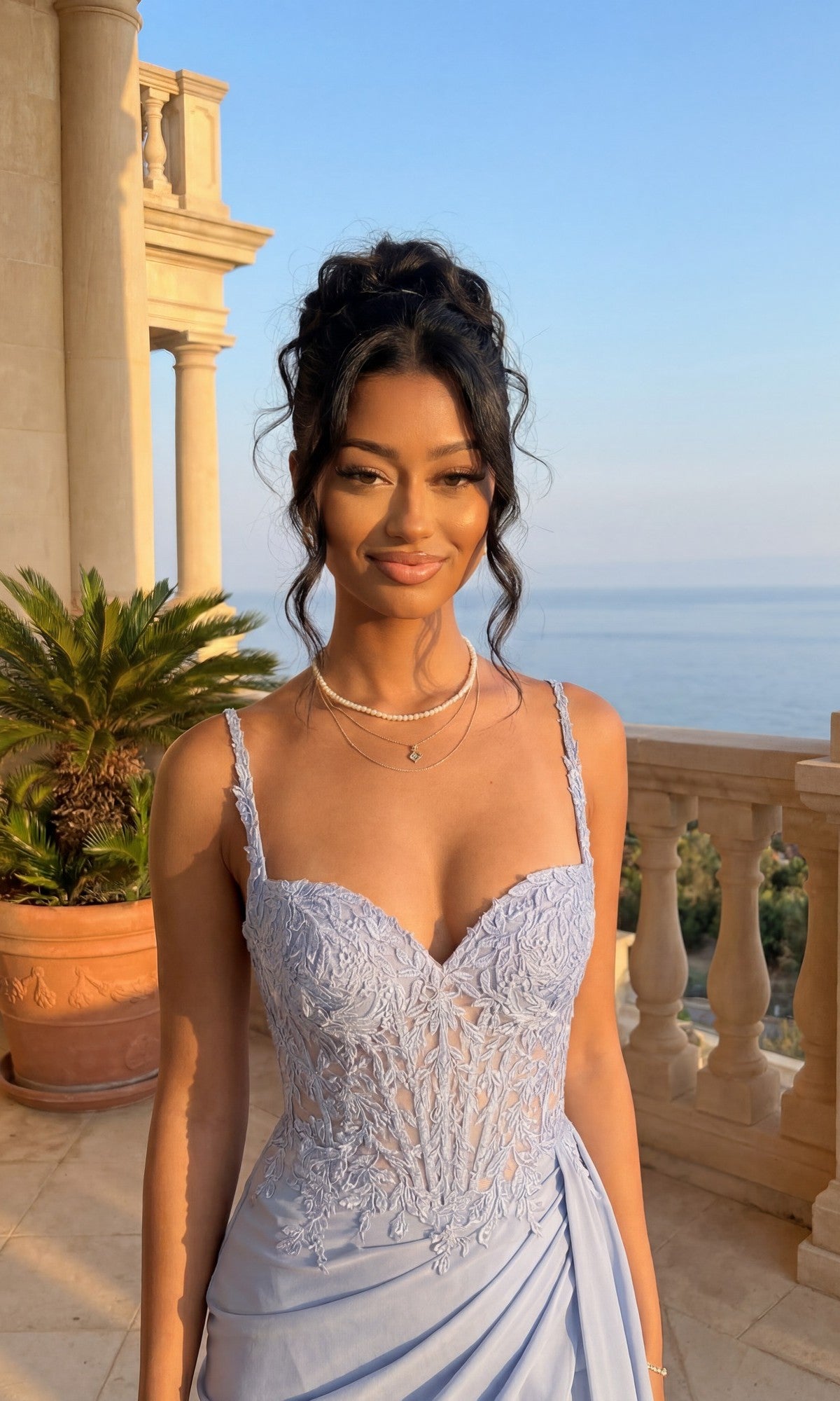 A woman in the Short Chiffon Hoco Dress: PromGirl Exclusive 250130 stands on a stone balcony with potted plants, overlooking the ocean on a sunny day.